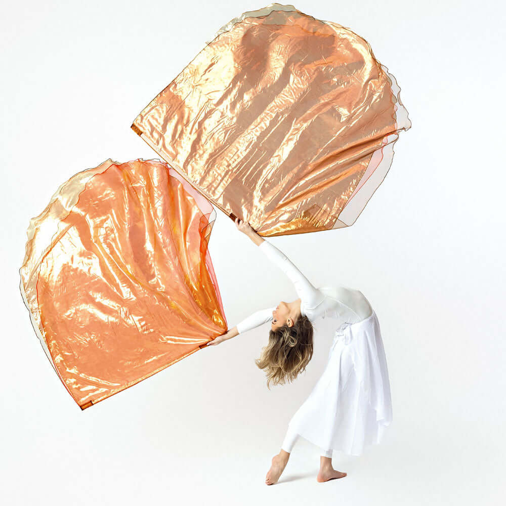A barefoot dancer dressed in white gracefully leans backward while holding two large, flowing worship flags in shades of bronze, gold, and soft red. The movement of the flags creates a dynamic arc against a plain white studio background.