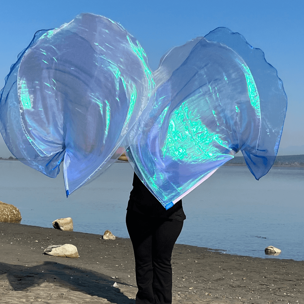 A pair of iridescent worship flags in shimmering light blue, lilac, and crystal hues billow in the air on a beach. The person holding the flags is mostly hidden behind the flowing fabric, creating a silhouette effect. The background shows calm ocean water and a clear blue sky.