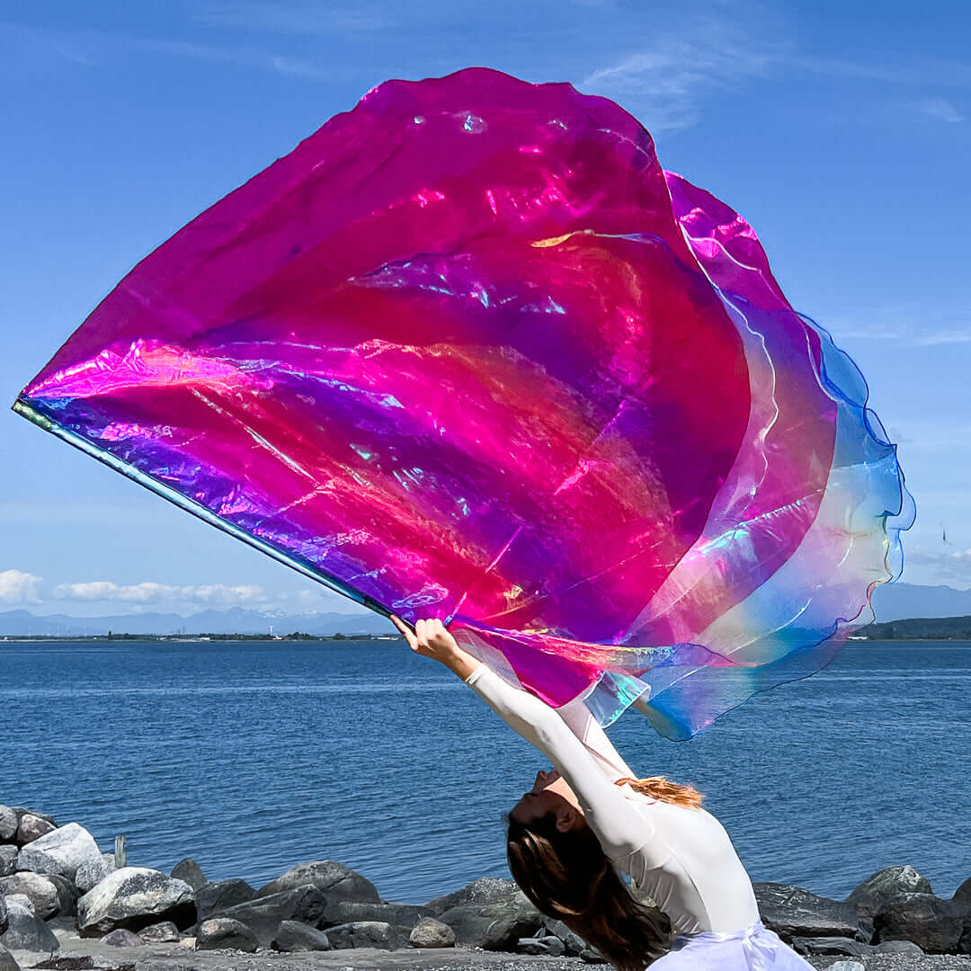 A worship dancer gracefully arches backward on a rocky shoreline, lifting a radiant worship flag in a dramatic movement. The flag features shimmering rainbow, pink, and crystal tones, with layers of translucent fabric catching the sunlight and casting prismatic reflections. Behind her, a tranquil sea and distant mountains enhance the sense of worship and freedom.