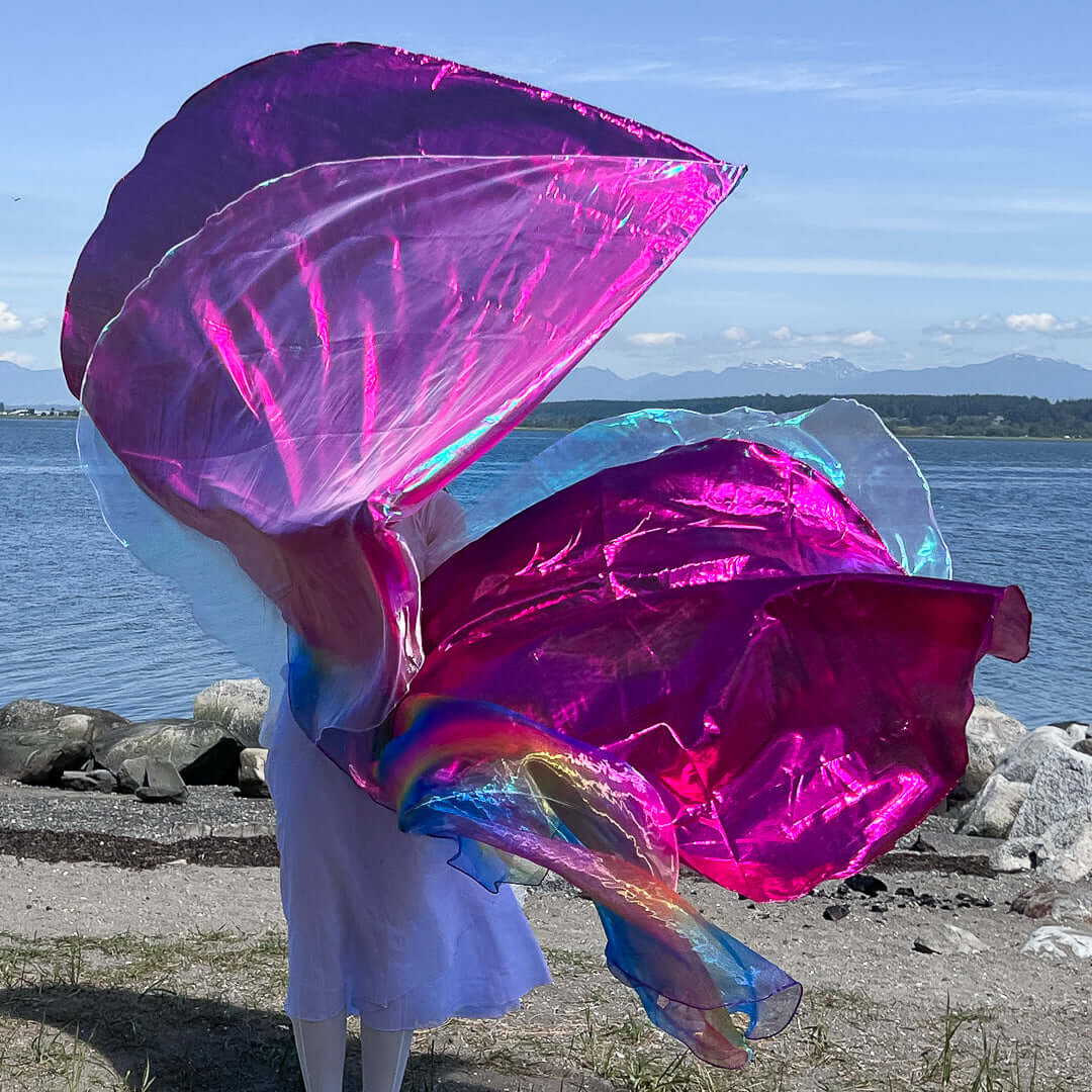 A worshipper in a flowing white dress holds two large, iridescent worship flags in shades of pink, rainbow, and crystal. The flags billow dramatically in the breeze, creating an energetic swirl of color. The scene is set on a rocky beach with a calm ocean and mountain range in the distance under a clear blue sky.