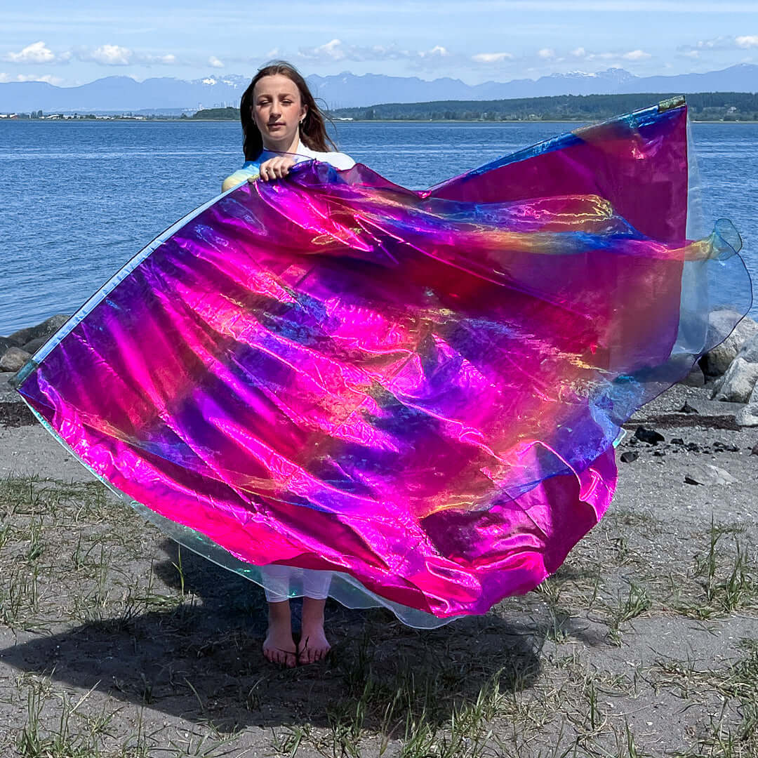 A woman stands barefoot on a beach holding a large, flowing worship flag made from iridescent fabric in vibrant shades of rainbow, pink, and crystal. The flag shimmers with dynamic hues of fuchsia, blue, and hints of gold and green, catching the sunlight as it ripples in the breeze. Behind her, a calm body of water and distant mountains create a peaceful and majestic backdrop.