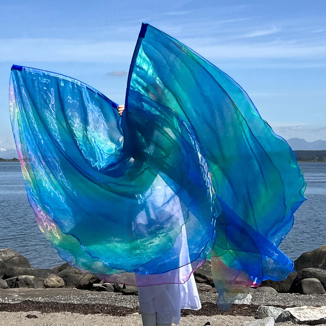 A dancer in a white outfit lifts two large worship flags at the waterfront. The flags shimmer in hues of blue, green, and iridescent rainbow, catching the sunlight and billowing dramatically against the sky and mountains in the background.