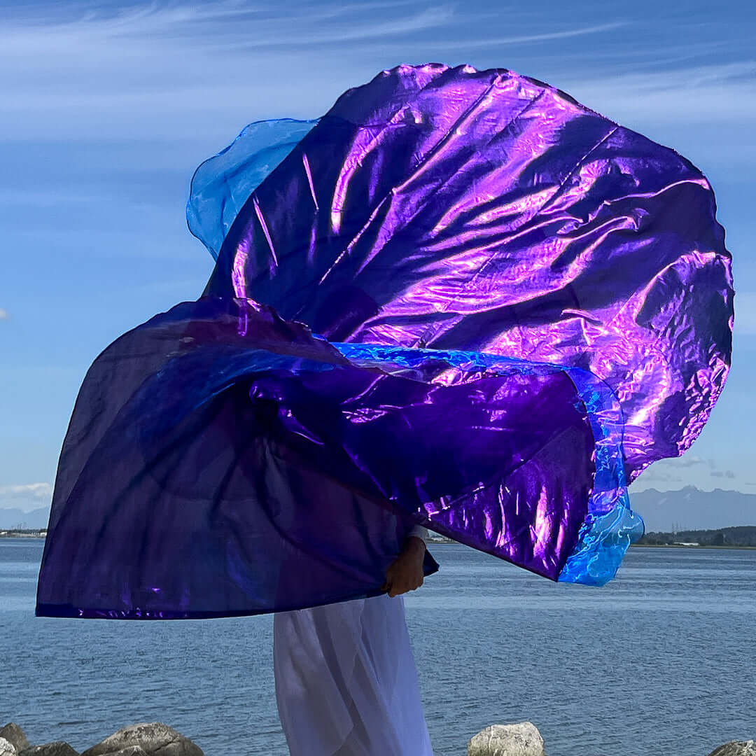 A dancer stands at the waterfront, enveloped by a dramatic swirl of worship flags. The flags are a rich, iridescent purple with electric blue edges, flowing outward in a wide arc. The person, mostly obscured by the movement of the flags, wears a white outfit, blending into the soft light of the shoreline. The scene is set against a tranquil lake and a clear, slightly clouded sky.