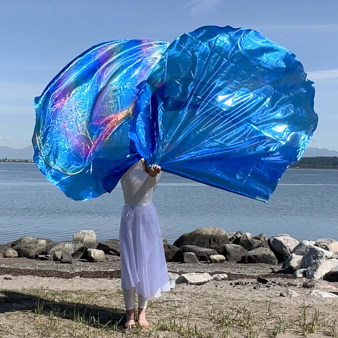 A woman dressed in white stands barefoot by the seashore, holding up two large worship flags. The flag on the left glows with iridescent rainbow tones, while the one on the right is a vibrant blue. The ocean, stones, and mountains form a peaceful natural backdrop under a clear sky.