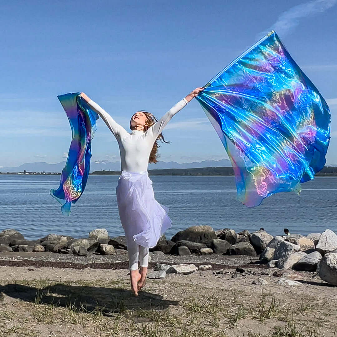 A barefoot woman dressed in white leaps joyfully into the air on a rocky beach, arms lifted high while holding two worship flags. The flags shimmer with radiant blue and rainbow hues, glowing against a backdrop of calm waters, distant mountains, and a clear blue sky.