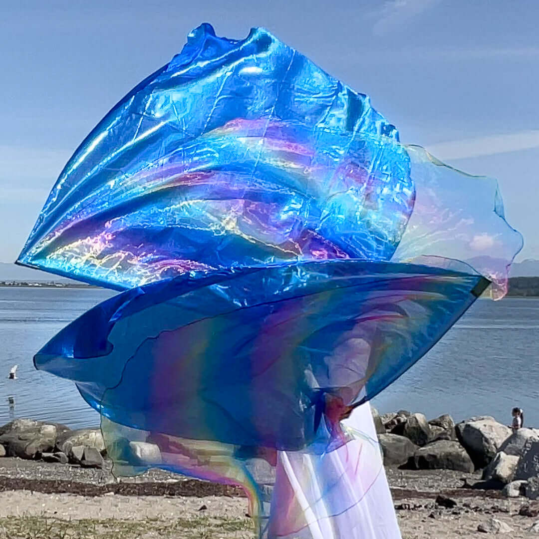 A worshipper dressed in white waves two vibrant flags along a peaceful shoreline. The flags shimmer with iridescent rainbow patterns and brilliant blue tones, catching the sunlight as they billow dramatically in the air. Behind them, still waters and a rocky beach create a serene backdrop.