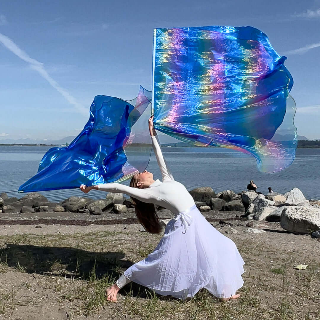 A worship dancer in a flowing white outfit performs a graceful backbend on a rocky beach, lifting two radiant worship flags. The flags shimmer in shades of electric blue with iridescent rainbow highlights, capturing the sunlight and reflecting the movement of praise against the calm water and clear sky.