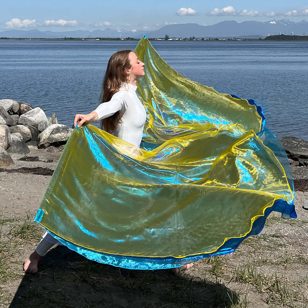 A worshipper in a white outfit gracefully moves with a vibrant pair of worship flags by the shoreline. The flags shimmer with translucent layers of yellow and blue, catching the sunlight. In the background, calm waters and distant mountains frame the peaceful and powerful moment of creative worship.
