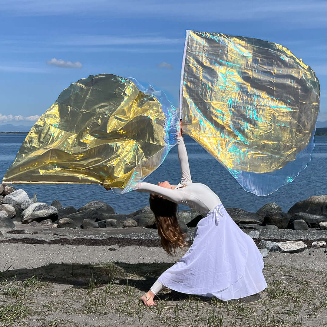 A worshipper dressed in white performs a dramatic backbend on a beach while lifting shimmering crystal and gold worship flags. The sunlight enhances the iridescence of the fabric, which arcs gracefully in midair, reflecting sky blue and golden hues. The water and mountains in the background provide a serene and majestic setting.