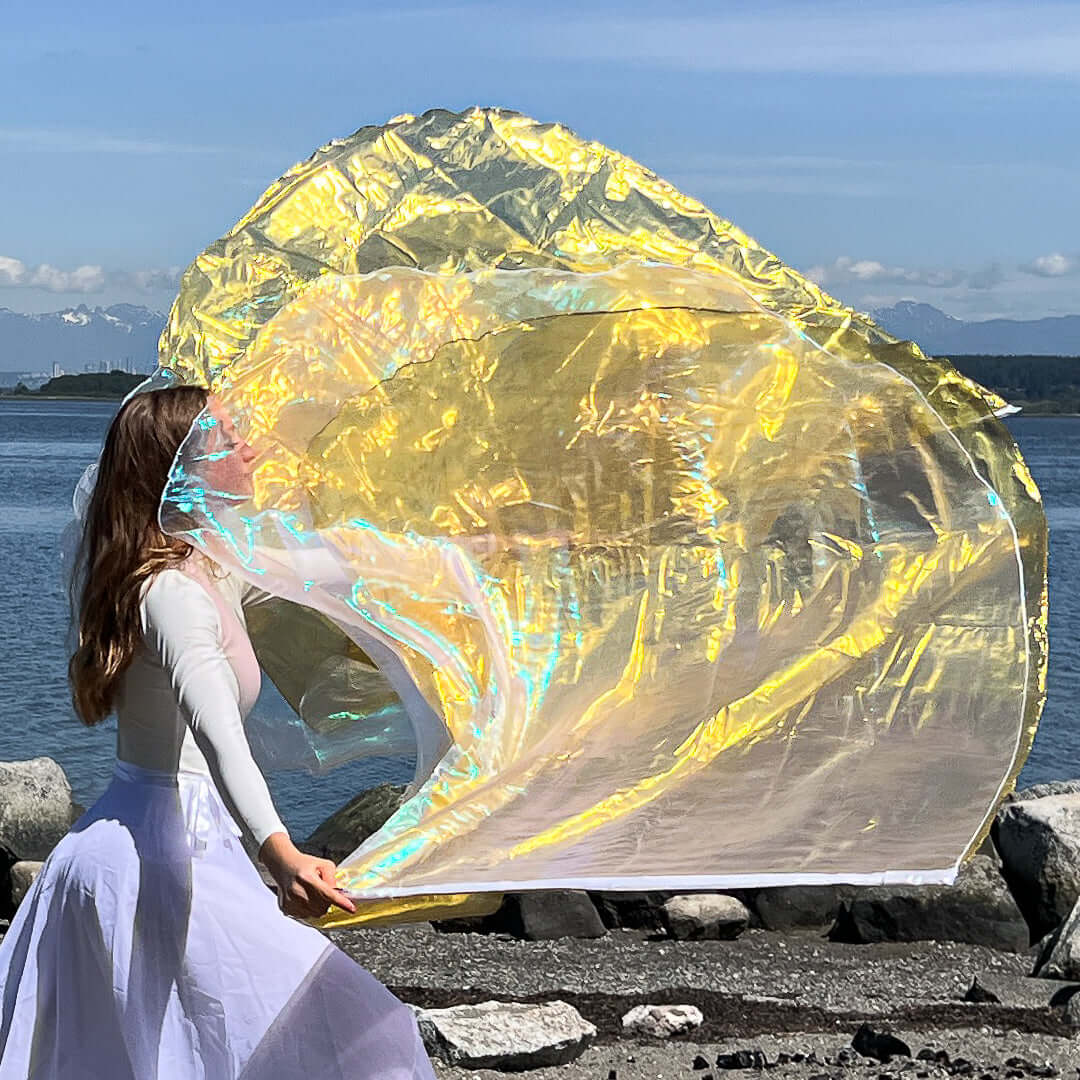 A woman dressed in a white worship outfit waves two large iridescent worship flags by the ocean. The flags shimmer with hues of crystal-clear white and vibrant gold, capturing the sunlight against a backdrop of calm water and distant mountains.