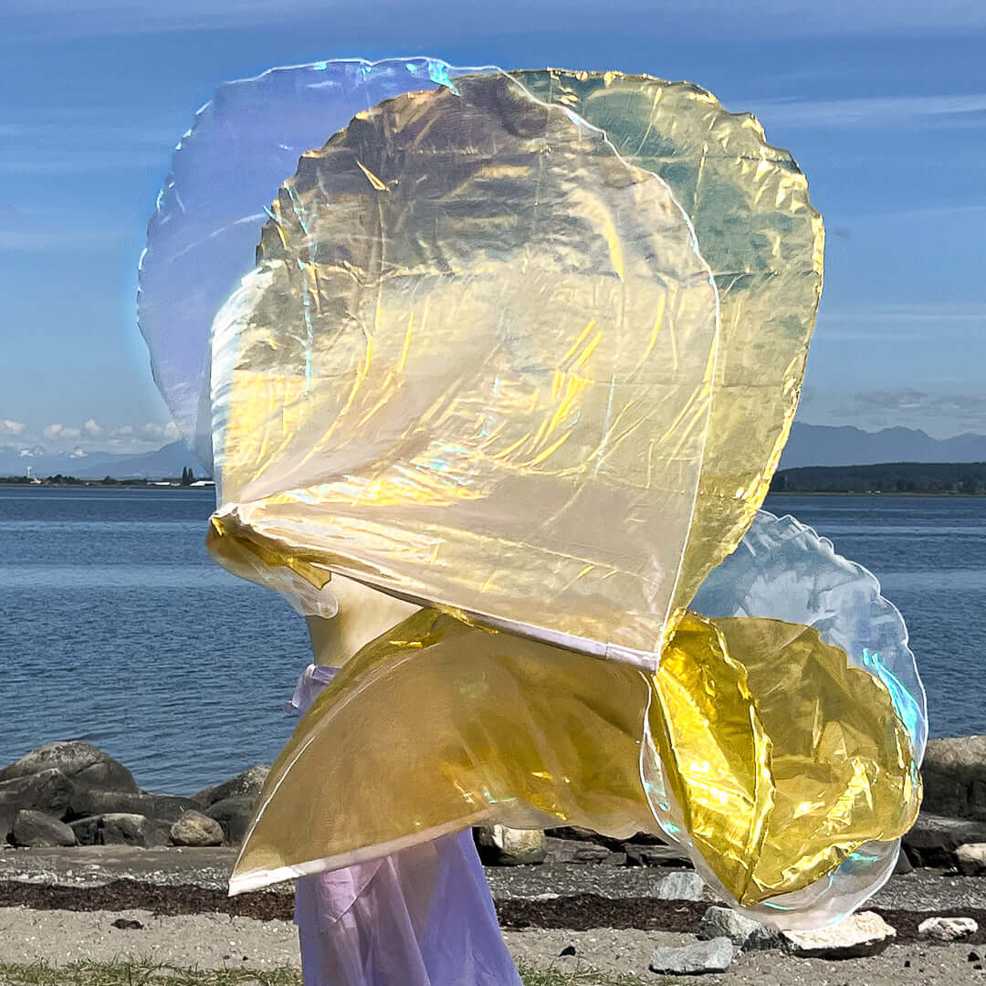 A person waves two layered worship flags by the seaside, with colors reflecting crystal, gold, and subtle lilac tones. The shimmering fabric glows under the sunlight, creating an ethereal, flame-like movement against a backdrop of water and distant mountains.