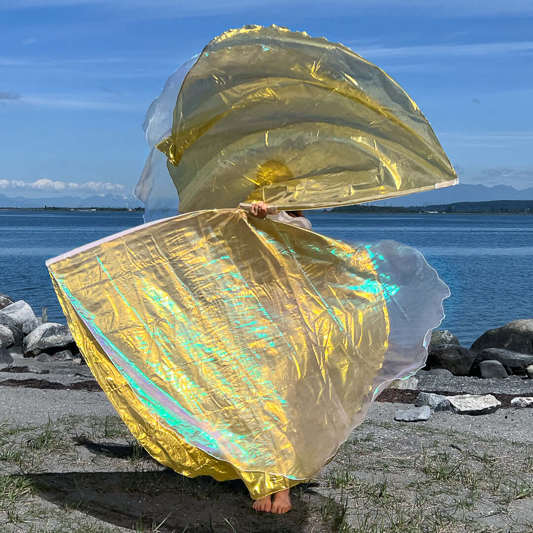A person worships at the water’s edge with large layered flags in crystal and gold. The iridescent fabric glows under the sunlight, creating a luminous, flowing effect that reflects the blue sky and lake. The movement of the flags forms graceful curves, capturing a moment of expressive praise.