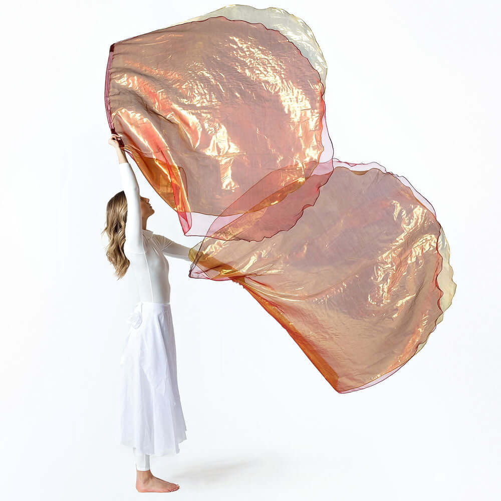 A worshipper dressed in white lifts a pair of flowing bronze and gold shimmer flags high into the air, creating a graceful arc against a clean white backdrop.