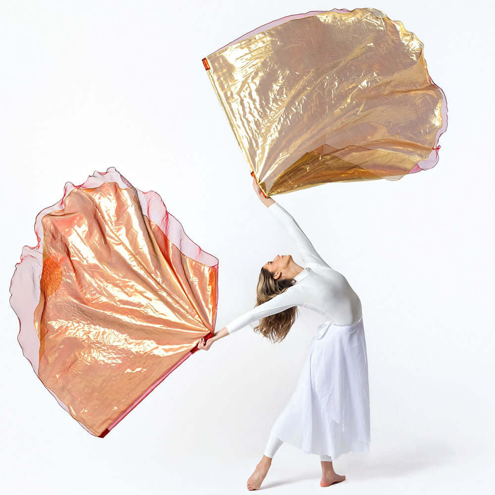 A worship dancer in a white outfit stretches gracefully, holding large flowing bronze and gold shimmer worship flags that billow like fire in motion against a clean white background.