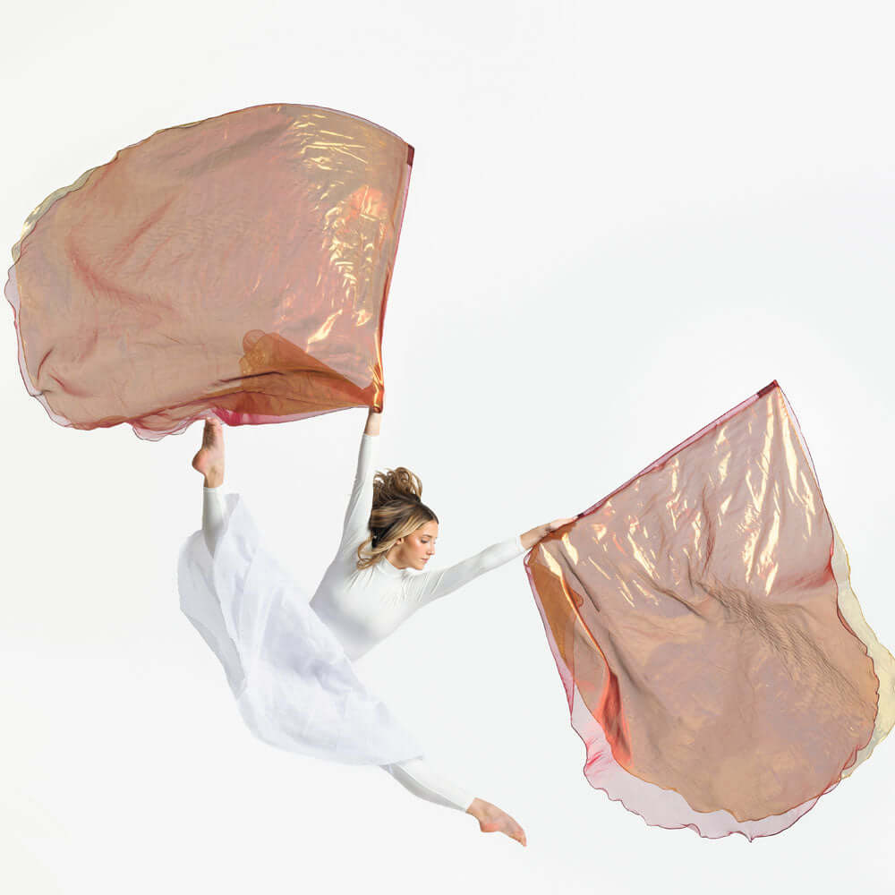 A dancer in a flowing white dress leaps mid-air, holding a pair of bronze and gold worship flags that shimmer with gold highlights, set against a clean white background.