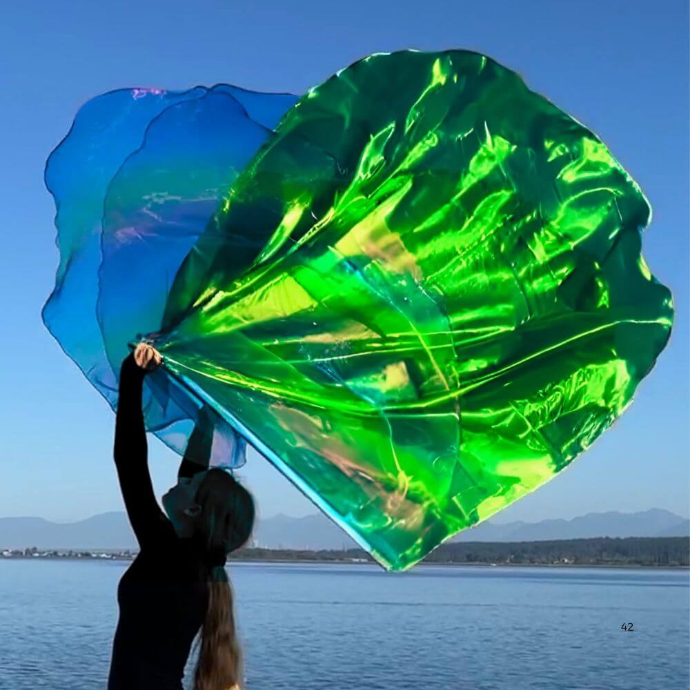 Woman worshipping with vibrant green and blue Jehovah Nissi prophetic worship flags by the water; angel wing flags for praise dance and spiritual warfare.