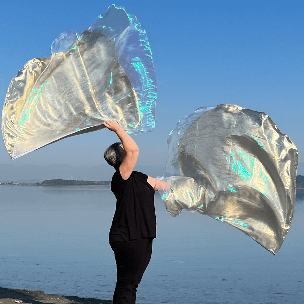 Woman worshipping with flowing iridescent lilac and gold flags at the water’s edge on a clear day.