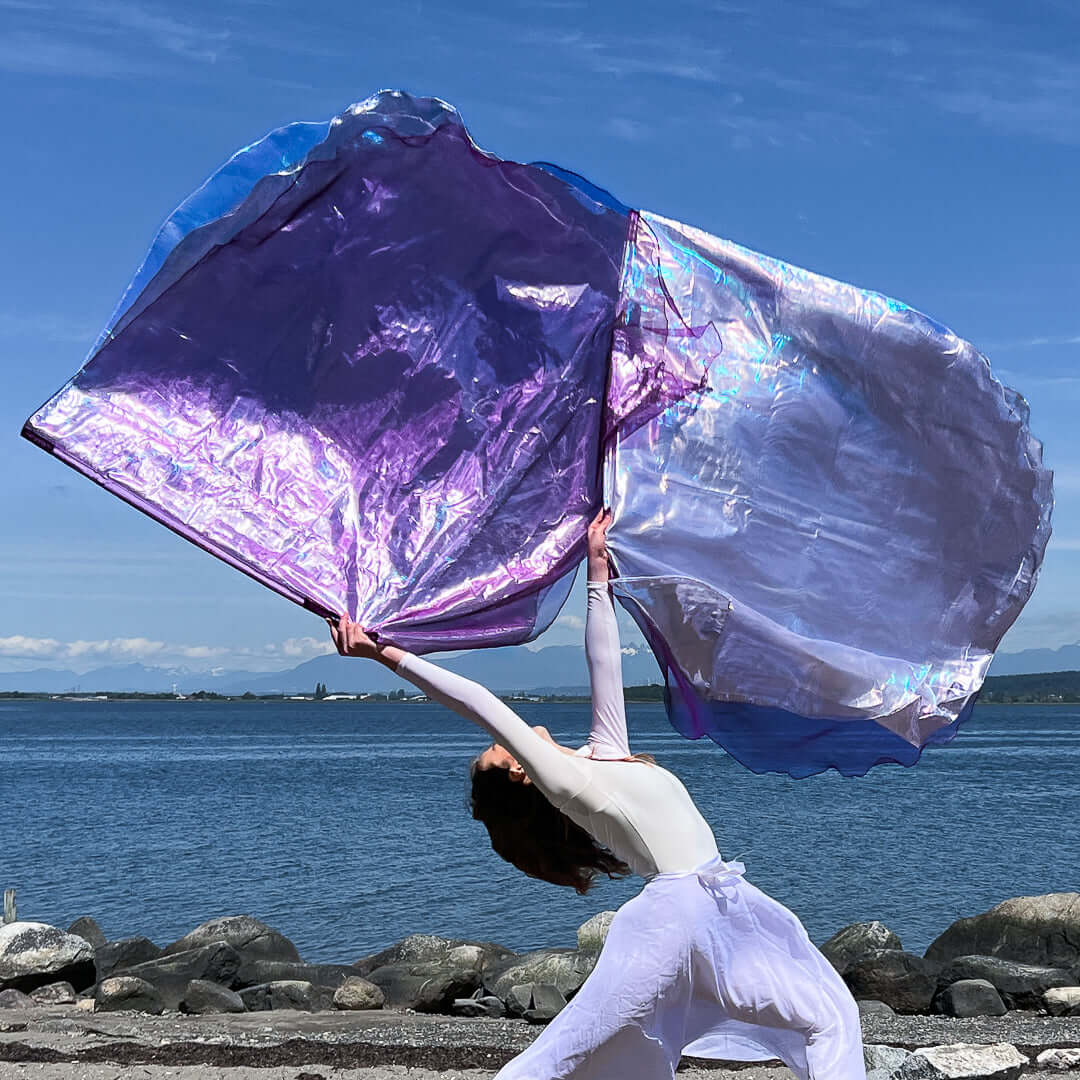 Woman in white worship attire dancing on a rocky shore with large flowing worship flags in shimmering purple, silver, and crystal tones.