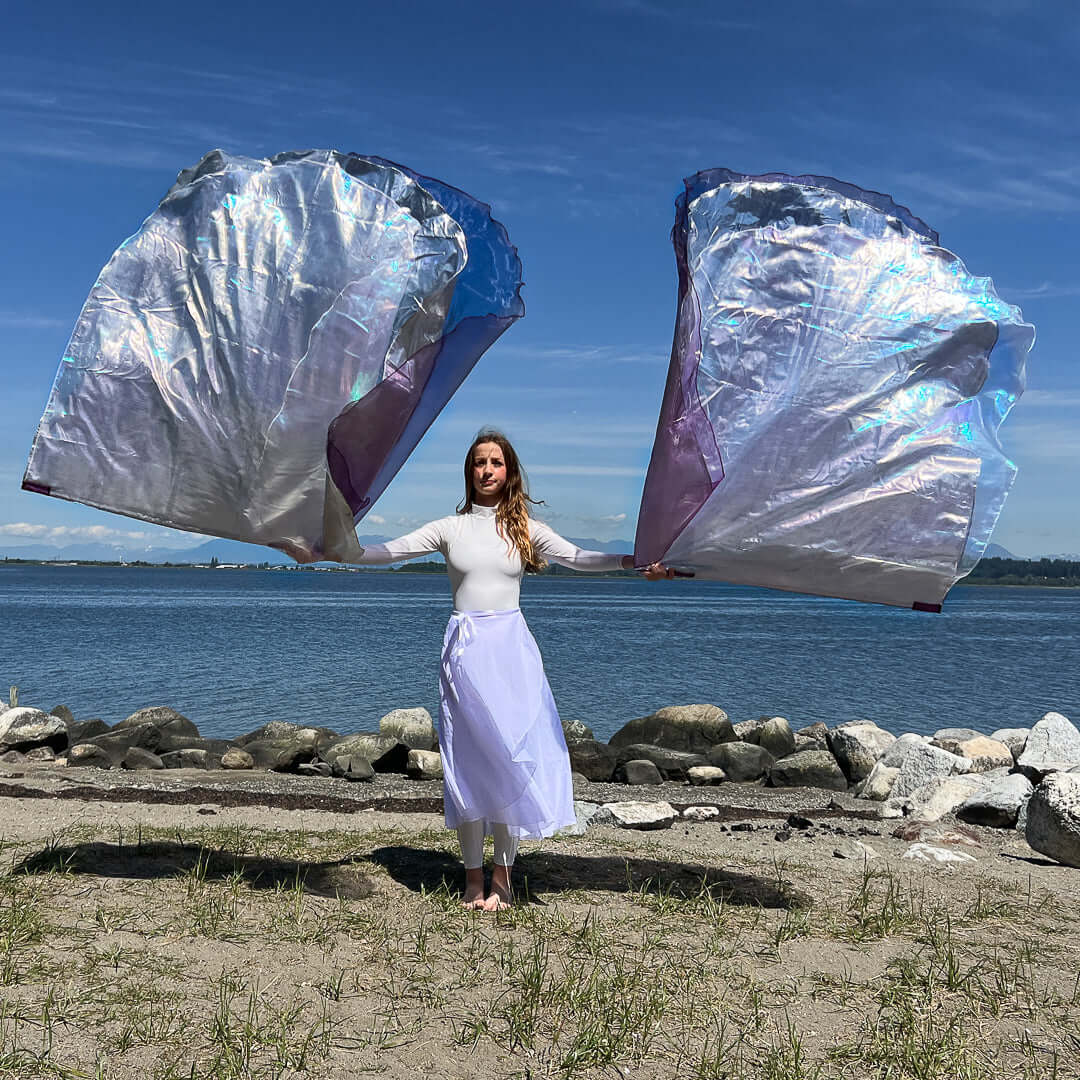 Woman in white praise garments standing with arms outstretched, holding large silver, purple, and crystal worship flags by the oceanfront.