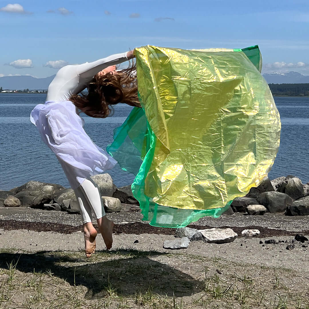 Woman in white worship attire mid-leap on a rocky shoreline, holding a large green and gold flag in an expressive act of praise dance.