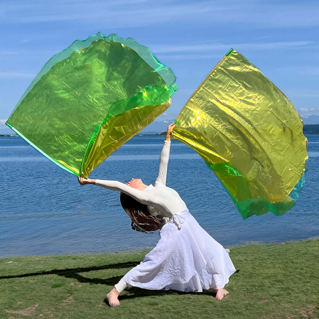 Woman in white worship attire dancing on grass near water, holding large green and gold worship flags in a dramatic pose of expressive praise.