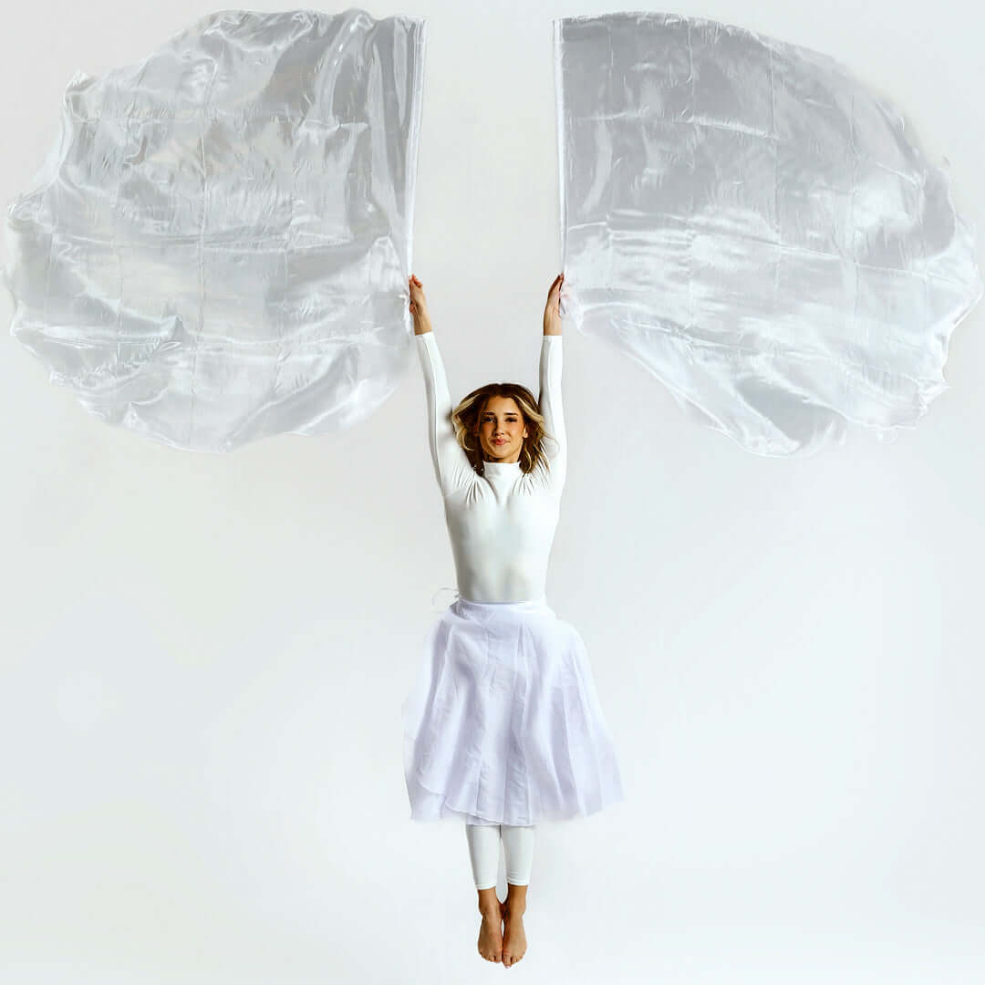 Woman in white worship attire leaping with both arms raised, holding large white worship flags that flow above her in a moment of vertical praise.