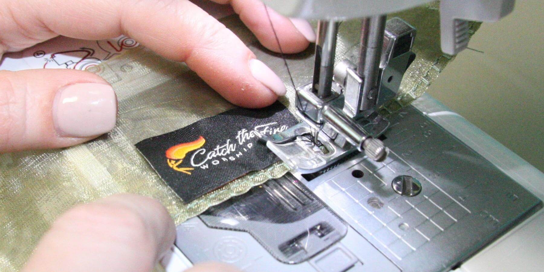 Close-up of hands sewing a Catch the Fire Worship Flags label onto a gold shimmer flag using a sewing machine, highlighting handcrafted quality and Christian artisan craftsmanship.