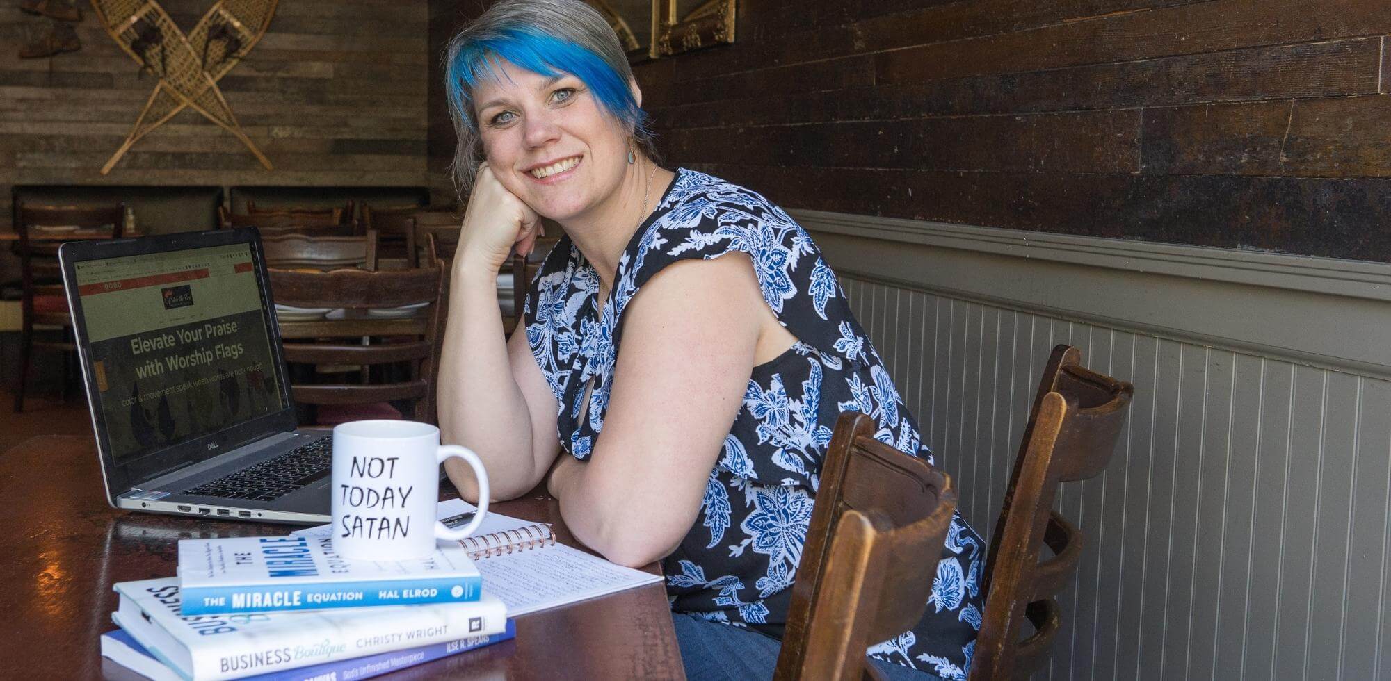 Woman with bright blue hair smiling while working on her laptop in a rustic café, with Catch the Fire Worship Flags website on screen, a “Not Today Satan” mug, and Christian business books nearby — symbolizing joyful spiritual entrepreneurship and faith-driven creativity.