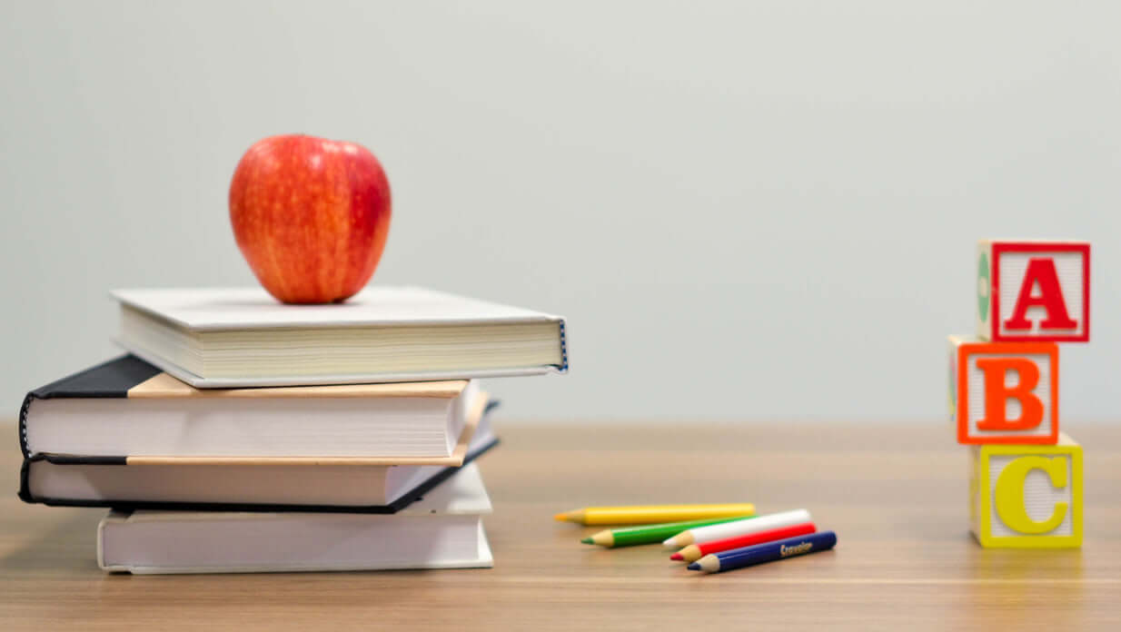 Stack of books with a red apple, colored pencils, and ABC letter blocks on a wooden desk — symbolizing foundational learning, creativity, and the heart of teaching for the Fire Catchers Classroom from Catch the Fire Worship Flags.