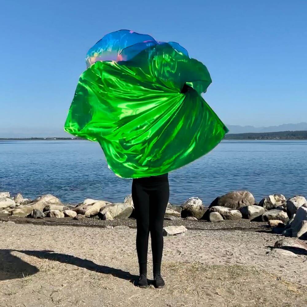 Worshipper waving large green and iridescent flags by the water, showcasing vibrant movement and outdoor praise setting.