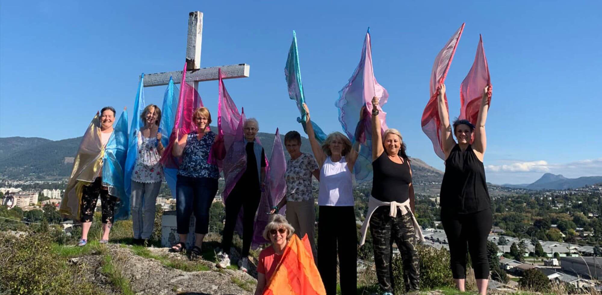 Group of worshippers waving colorful worship flags on a mountaintop beside a wooden cross, declaring God's glory over the city below. A joyful prophetic act of intercession and unity through Catch the Fire Worship Flags.