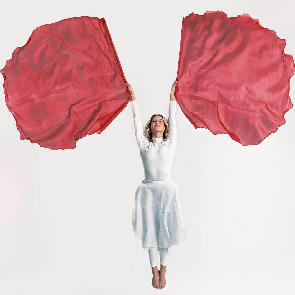 Worship dancer in white garment jumps gracefully while holding red praise flags, symbolizing spiritual warfare and the victory of Christ, against a white studio backdrop.