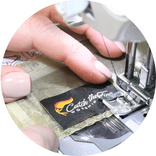 Close-up of hands sewing a Catch the Fire Worship Flags label onto a olive green sheer flag using a sewing machine, highlighting handcrafted quality and Christian artisan craftsmanship.