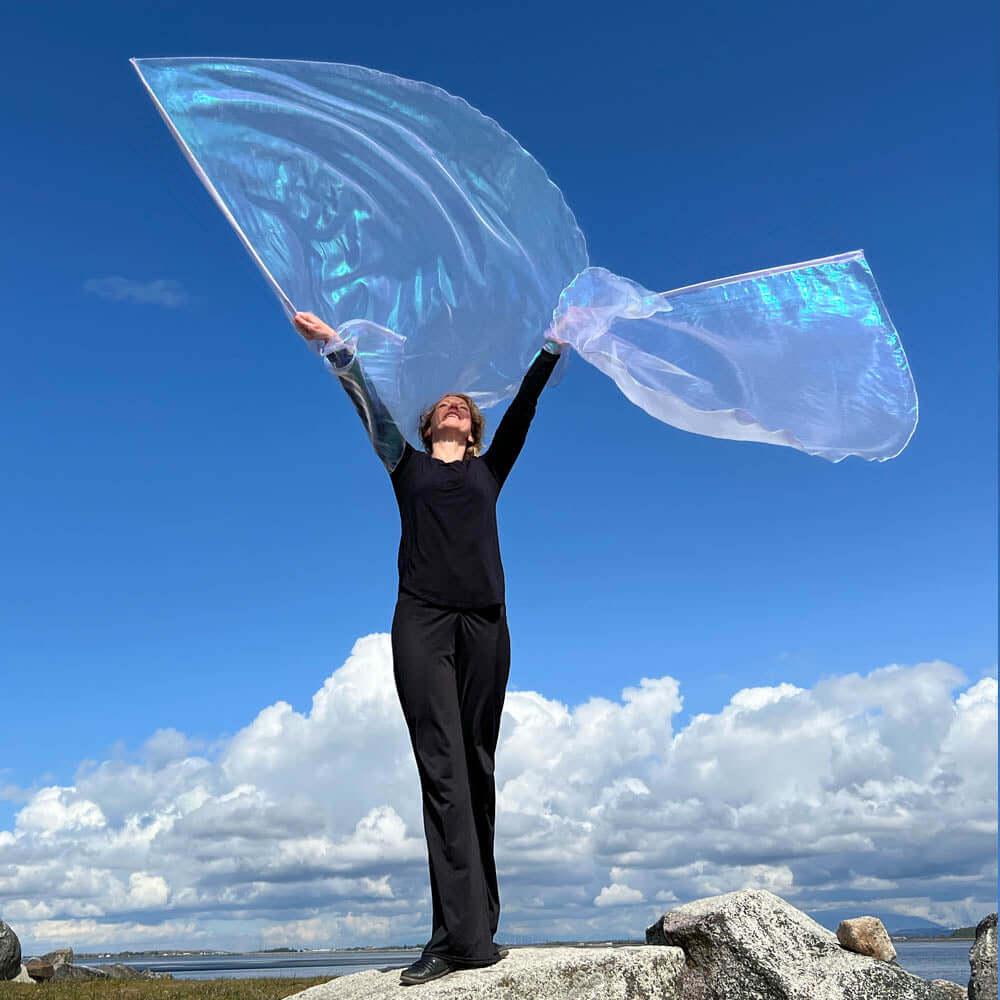 Woman worshipping with large iridescent lilac-colored flags billowing against a bright blue sky, demonstrating expressive movement in prophetic praise dance.