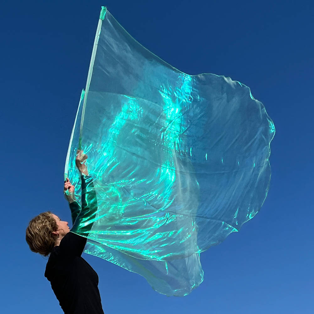 Worshipper holding a large, flowing seaglass-colored worship flag made of iridescent sheer fabric, glowing with aqua and turquoise hues under a vibrant blue sky.