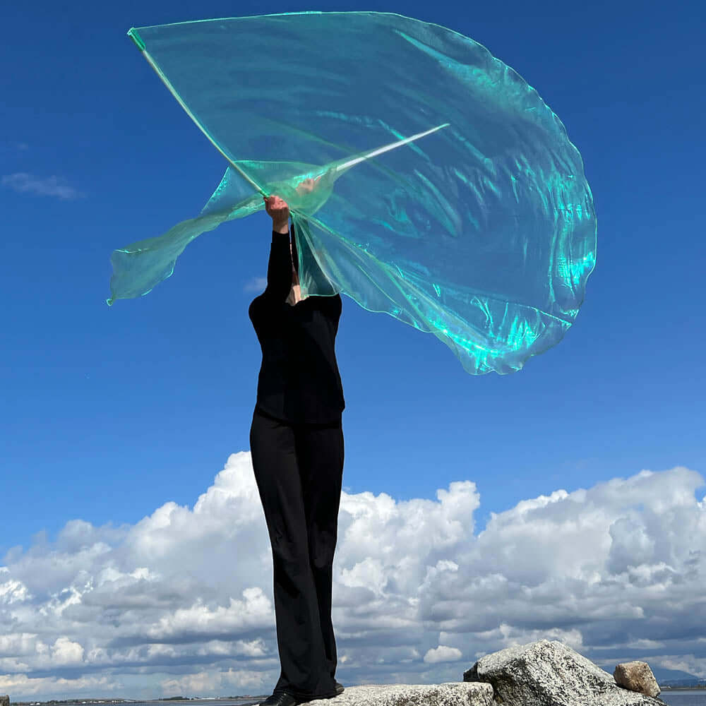 Worship dancer standing on a rocky ledge, lifting a large iridescent seaglass-colored worship flag against a dramatic sky with white clouds and deep blue background.
