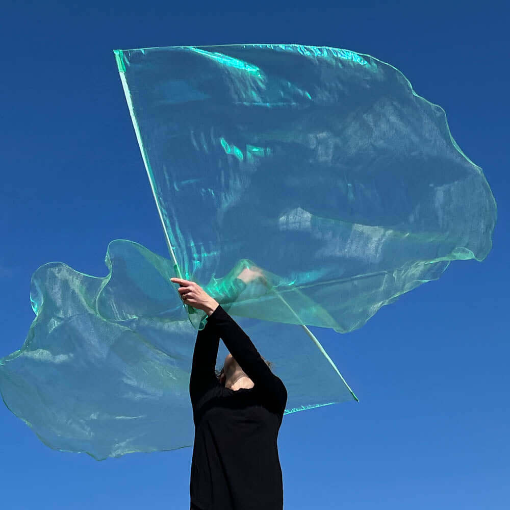 Worshipper dressed in black gracefully lifting a translucent seaglass-colored worship flag under a vivid blue sky, with both arms extended upward.