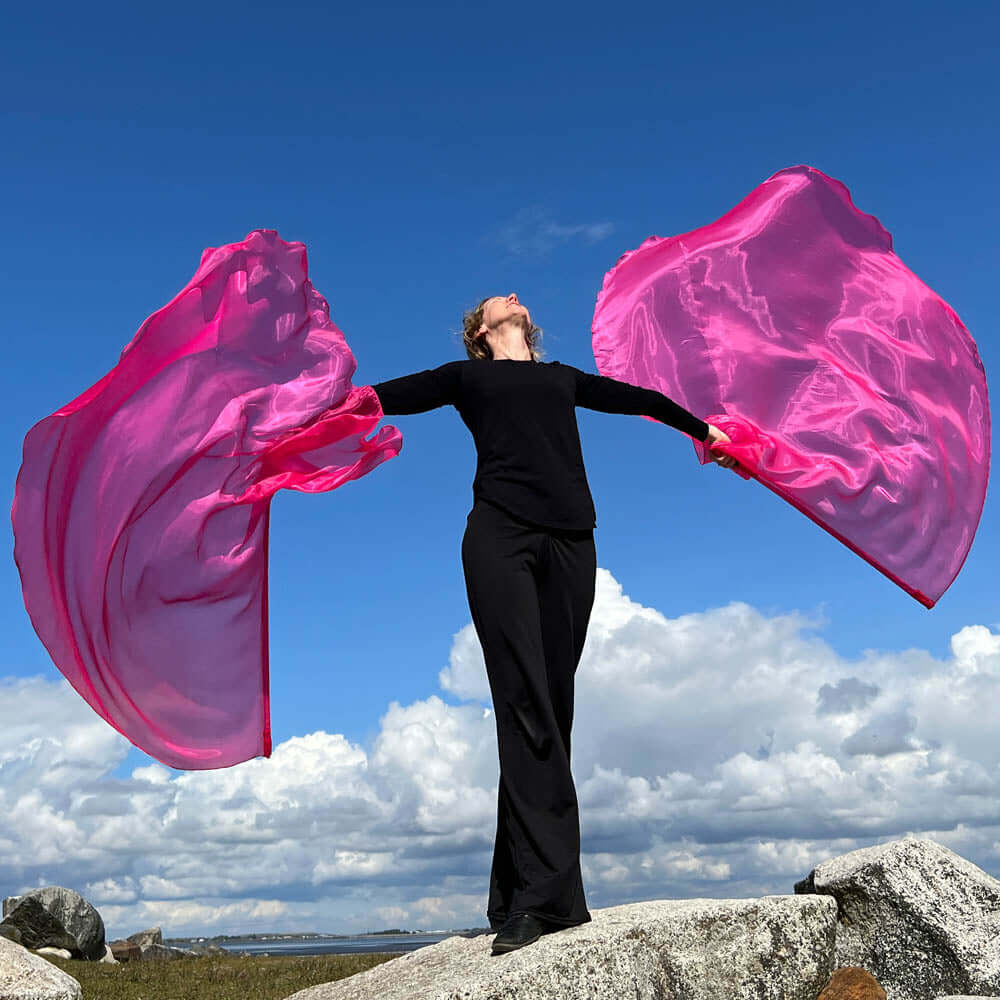 Woman worshipping outdoors with hot pink worship flags, arms raised in praise on a large rock under a bright blue sky with clouds.