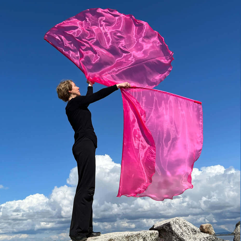 Praise dancer worshipping with hot pink worship flags, standing on a rock against a blue sky and clouds — expressive Christian worship outdoors.