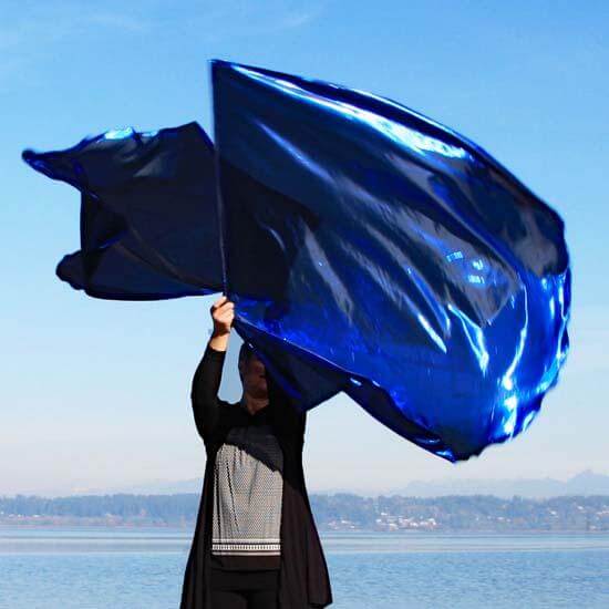 Worshipper lifting vibrant deep blue metallic worship flags at the water's edge, flags billowing in the wind under a clear blue sky.