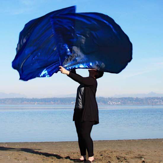 Worshipper waving majestic sapphire blue metallic worship flags on a sandy shoreline, under a radiant blue sky with distant mountains in view.