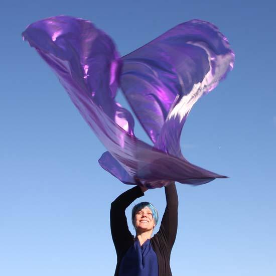 Joyful woman worshipping with vibrant purple shimmer flags against a clear blue sky, lifting them high in expressive praise.