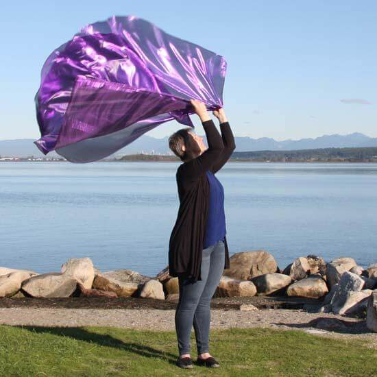 Woman worshipping with large, shimmering purple worship flags at the waterfront, standing on green grass with a view of mountains and blue sky.