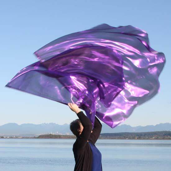 Worship dancer lifting vibrant purple flags in the air beside a peaceful lake with distant mountains under a clear sky.