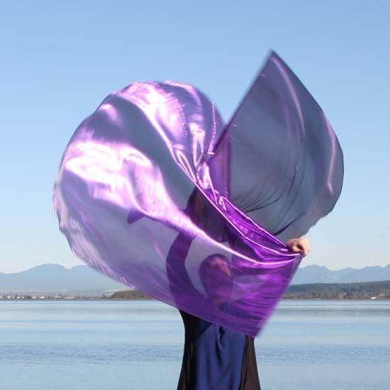 Worshipper holding up large flowing purple worship flags beside a calm lake under clear blue skies, with mountains visible in the background.