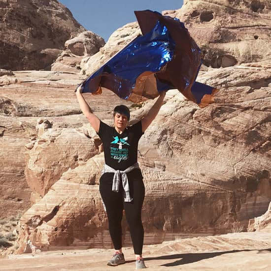 Woman lifting worship flags in bronze and royal blue while standing on desert rock formations, with sunlight highlighting the shimmer of the fabric.