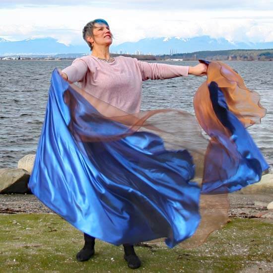 Woman worshipping near a lake with large multi-layer worship flags in bronze and royal blue, flowing dramatically in the wind against a scenic mountain backdrop.