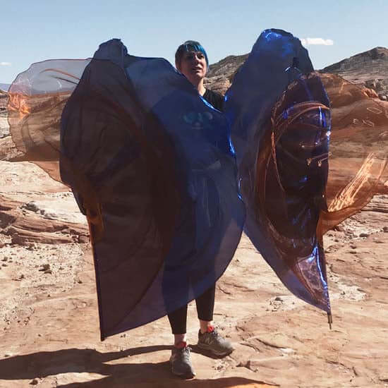 Woman lifting worship flags in bronze and royal blue while standing on desert rock formations, with sunlight highlighting the shimmer of the fabric.