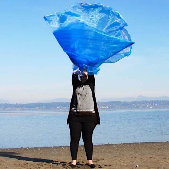 Person standing on a sandy shoreline, lifting sheer blue and white worship flags into the air with both hands, with a lake and distant mountains in the background.