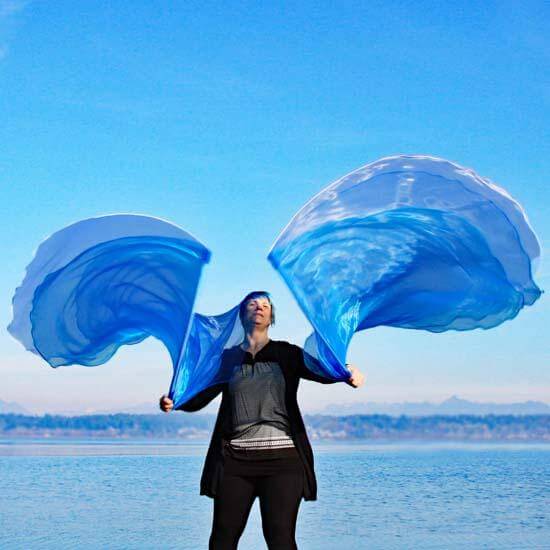 Woman worshipping outdoors with sheer blue and white worship flags, arms outstretched as the flags flow upward against a clear blue sky and lake backdrop.